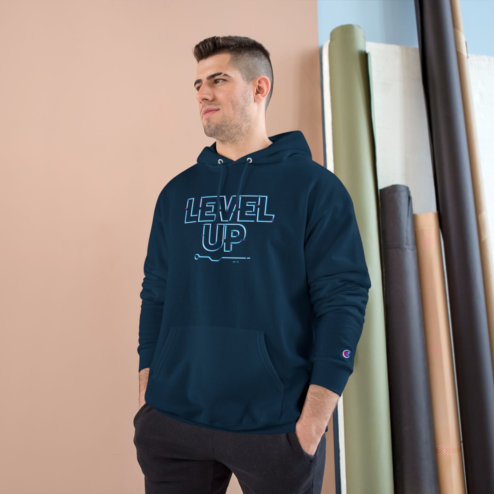 Man wearing a navy hoodie with 'LEVEL UP' text, standing against a beige wall with fabric rolls.