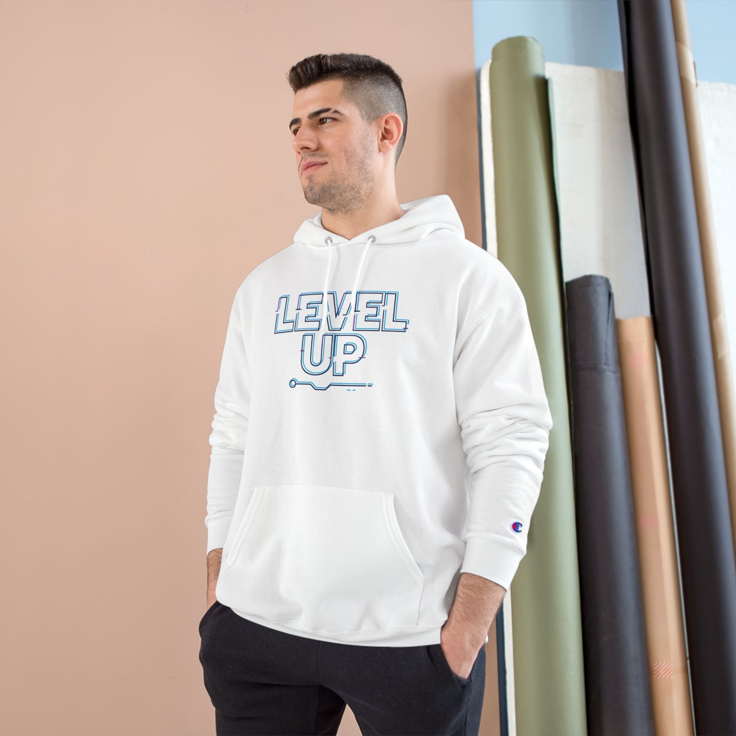 Man wearing a white hoodie with 'LEVEL UP' text, standing against a beige wall with rolled-up rugs.
