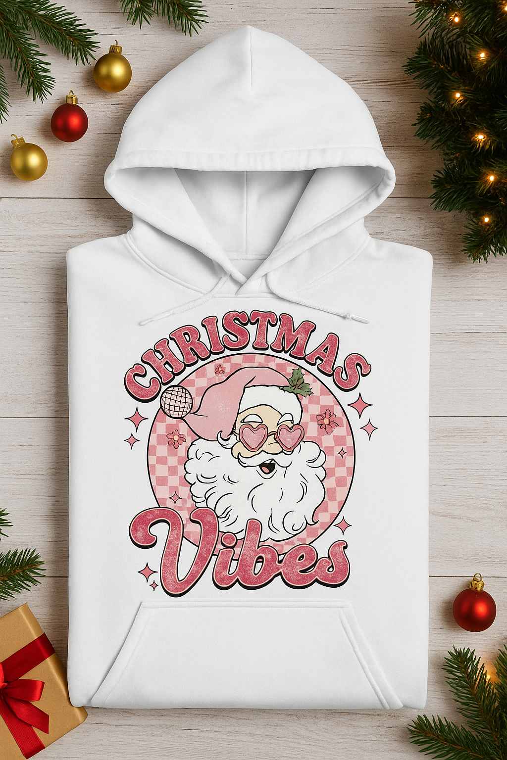 White hoodie with 'Christmas Vibes' design featuring Santa Claus, surrounded by Christmas decorations on a wooden surface.