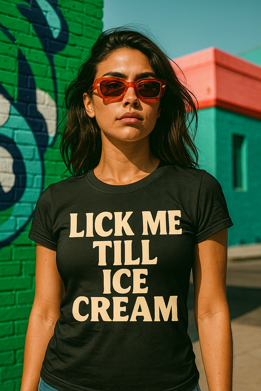A young woman wearing a fitted Black t-shirt with the bold cream white text “LICK ME TILL ICE CREAM” printed on the front, standing against a graphitic background.