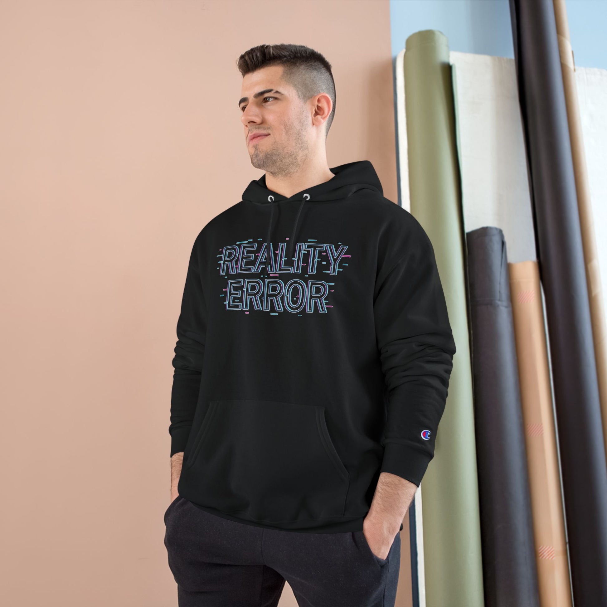 Man wearing a black hoodie with 'REALITY ERROR' text, standing against a beige wall with rolled-up rugs.